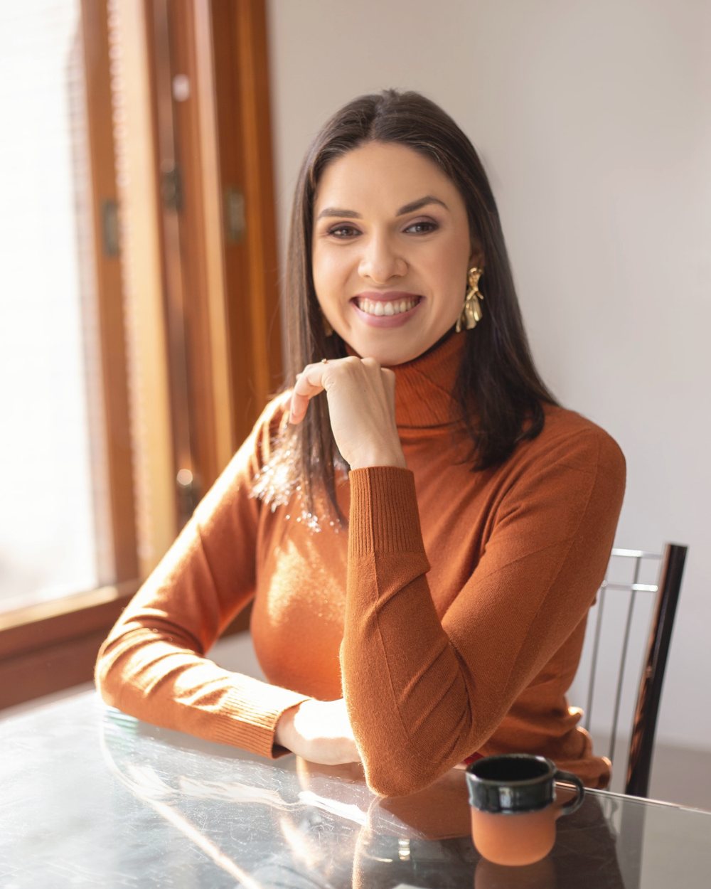 Daiana Prestes sorrindo, sentada à mesa perto da janela